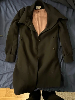 Svart kappa med bred krage och knappar - Sjukt snygg svart kappa med bred krage och snygga knappar framtill. Kragen går att stänga. Figurnära passform och helsvart utsida, med ljusrosa foder på insidan. Långa ärmar och stilren design som ger en clean look. Perfekt för dig som gillar klassiska och minimalistiska plagg.