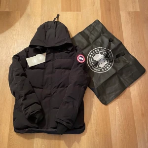 Svart dunjacka från Canada Goose - Svart dunjacka från Canada Goose med huva och klassisk logotyp på ärmen. Jackan har en stilren, quiltad design och ribbade muddar vid ärmslut. Perfekt för kalla dagar och har en snygg, enkel look som funkar till det mesta.