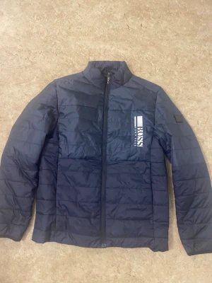 Mörkblå pufferjacka från Hugo Boss - Snygg mörkblå pufferjacka från Hugo Boss med diskret logga på bröstet. Jackan har quiltad design, dragkedja framtill och hög krage. Perfekt för kyliga dagar och har en clean, modern look som funkar till många stilar.