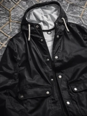 Svart vindjacka med huva och snörning - Svart jacka i slitstarkt material med huva och praktiska fickor. Varm och bekväm med stilren design – perfekt för vardagsbruk under kyliga dagar. Fint skick.