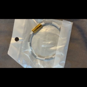 Två armband med guldfärgat lås - Säljer två coola armband med cylinderformat guldfärgat lås. Ett är vitt med svarta detaljer och det andra är helsvart. Båda har ett sportigt repmönster och känns riktigt trendiga att ha på handleden.