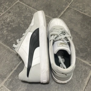 Vita och svarta sneakers från Puma - Snygga vita sneakers från Puma med klassisk svart sidodetalj och guldfärgad Puma-logga på plösen. Skorna har snörning, rund tå och platt sula. Ovandelen är i skinnliknande material och insidan är vadderad för extra komfort. Perfekta för en sportig och stilren look.