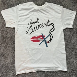 Saint Laurent No Smoking T shirt - Vit t-shirt från Saint Laurent med coolt grafiskt tryck på framsidan. Trycket består av röda läppar, svarta och blå linjer samt texten 'Saint Laurent' i svart. Klassisk rund hals och korta ärmar. Perfekt för dig som gillar statement-plagg. Size L kan passa M