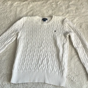 Vit kabelstickad tröja Polo Ralph Lauren - Vit kabelstickad tröja från Polo Ralph Lauren med klassisk rund halsringning och diskret broderad logga på bröstet. Tröjan har långa ärmar och ribbade muddar för en snygg passform. Perfekt för dig som gillar stilren och tidlös design.