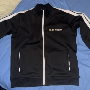 Palm Angels svart track jacket - Svart track jacket från Palm Angels med vita ränder längs ärmarna och vit dragkedja. Klassisk logga på bröstet och två fickor med dragkedja. Tillverkad i polyester för en sportig vibe och perfekt passform.  Det finns ett litet hål i den men jag har sytt den o den syns knappast när man har den på sig, produkten är inte äk*a