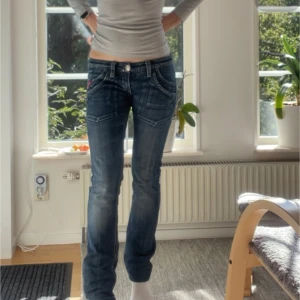 Blå bootcut jeans från My God - Säljer ett par blå jeans från My God med bootcut passform och låg midja. Storlek w27l34. Jeansen har coola detaljer som kontrastsömmar, slitningar och en röd detalj vid fickan. Klassisk femficksmodell med patch bak och dragkedja i gylfen. Perfekta för dig som gillar retrovibe. (Säljer dom för billigare på Vinted, olssonnelly)