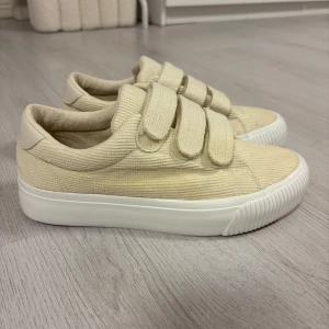 Beigea sneakers  - Snygga beige sneakers i manchestertyg med breda kardborreband och vit platt sula. Ser gulare ut på bild än i verkligheten.