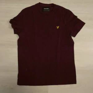 Vinröd t-shirt från Lyle & Scott - Säljer en vinröd t-shirt från Lyle & Scott med klassisk gul logga på bröstet. T-shirten har rund halsringning och korta ärmar. Skön och enkel modell i bomull som passar perfekt till jeans eller shorts.