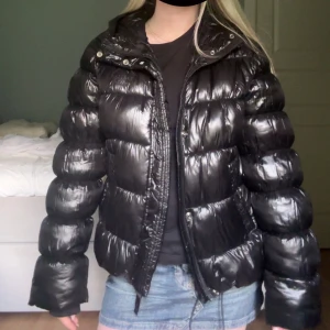 Svart glansig pufferjacka Superdry - Säljer en svart, glansig pufferjacka med huva och dragkedja. Jackan funkar som både höst och vinter jacka. Från Superdry.
