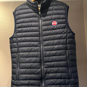 Mörkblå dunväst från Colmar - Snygg mörkblå dunväst från Colmar med quiltad design och dragkedja framtill. Västen har två praktiska fickor med dragkedja och en hög krage. Colmar-logga på bröstet och bak. Västen har även en ficka på insidan av västen. Finns även en liten defekt men inget som syns vid användning