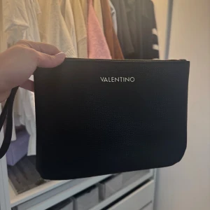 Svart kuvertväska från Valentino - Stilren svart kuvertväska från Valentino med diskret struktur i materialet och guldfärgad logga framtill. Väskan har dragkedja upptill och en smidig handledsrem, perfekt för att bära det viktigaste. Enkel och snygg design som passar till många outfits. Helt ny