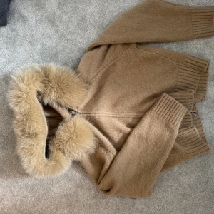 Beige stickad jacka med pälskrage från ALESSA - Snygg beige stickad jacka med fluffig pälskrage och dragkedja framtill. Jackan har ribbade muddar vid ärmslut och nederkant, perfekt för kyliga dagar. Ger en lyxig och mysig vibe till din outfit.
