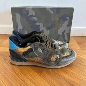 Valentino Rockrunner sneakers camo - Snygga Valentino Rockrunner sneakers med camouflage-mönster i grönt, brunt och svart. Skorna har blå detalj på hälen, snörning och coola nitar på sulan. Materialmix av skinn, mocka och textil ger en lyxig känsla 