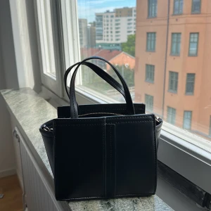 ZARA väska  - Snygg svart handväska i skinn med stilren, boxig form. Väskan har både korta handtag och två olika axelremmar – en bred textilrem och en cool guldfärgad kedja med skinn-detalj. Dragkedja upptill och minimalistisk design, perfekt för att lyfta din outfit.