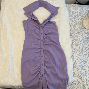 Lila ribbad kortklänning med knappar - Supersöt lila kortklänning i ribbat material med knappar hela vägen framtill och snygg rynkdetalj. Klänningen har en unik krage och är ärmlös, perfekt för varma dagar eller när du vill sticka ut lite extra.