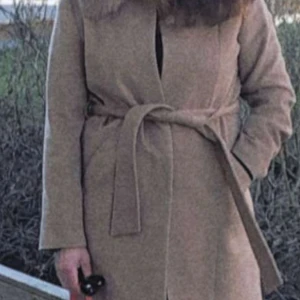 Beige kappa med pälsdetalj och bälte - Snygg beige kappa med bred krage i pälsimitation och knytbälte i midjan. Jackan har klassisk rak passform, två fickor fram och långa ärmar. Perfekt för dig som gillar stilrena och tidlösa plagg med en lyxig touch.