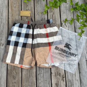 Burberry rutiga badshorts med snörning - Snygga badshorts från Burberry med klassiskt rutigt mönster i beige, svart, vit, grå och röd. Shortsen har elastisk midja med svart snörning och meshfoder. Bakficka med rutigt lock. Perfekt för sommaren och strandhäng.