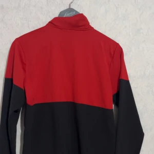 Röd och svart vindjacka Under Armour - Snygg röd och svart vindjacka från Under Armour med hel dragkedja framtill och två fickor med dragkedja. Jackan har hög krage och sportig look, perfekt för dig som gillar att sticka ut. Logga på bröstet och skön passform.