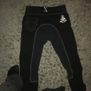 Svarta ridbyxor med vita sömmar - Svarta ridbyxor med vita kontrastsömmar och detaljer som broderad logga och flagga på fickan. Byxorna har förstärkning på insidan av benen och bälteshällor. Snygg passform och stretchigt material som ger bra rörelsefrihet. Märke HV polo