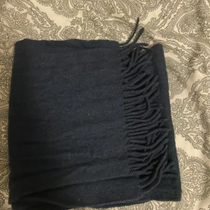 Mörkblå ullhalsduk med fransar - Snygg och klassisk mörkblå halsduk i mjuk ull med stilrena fransar längs ena kanten. Perfekt att svepa runt halsen för extra värme och en clean look. Passar dig som gillar enkel och tidlös stil.