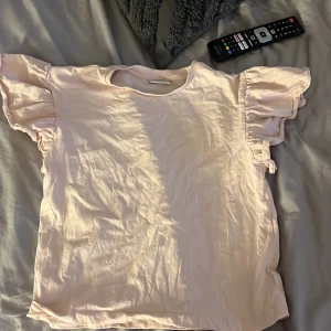 Ljusrosa volangtopp från Kappahl - Supersöt ljusrosa topp från Kappahl med volangiga korta ärmar som ger en extra touch. Tillverkad i mjuk bomull och perfekt för dig som gillar en enkel men ändå söt stil. Rund halsringning och loose fit för en chill vibe.