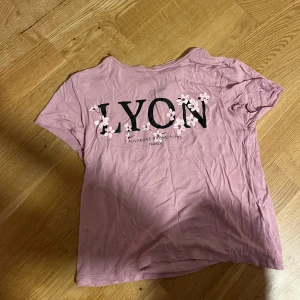 Rosa t-shirt med blommor och tryck - Snygg rosa t-shirt från HM i storlek XS med trycket 'LYON' och Auvergne-Rhône-Alpes, France på bröstet. Dekorerad med vita och rosa blommor över bokstäverna. Mjuk bomullskvalitet och klassisk rund hals. Perfekt för dig som gillar franska vibes och blommiga detaljer.