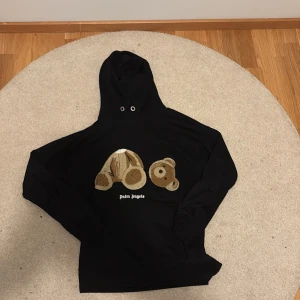 Svart Palm Angels hoodie med björn - Svart hoodie från Palm Angels med ikoniskt björn-motiv i beige och brunt framtill. Tröjan har huva med snörning och klassisk logga under motivet. Perfekt statement-plagg för dig som gillar streetwear och unika detaljer.