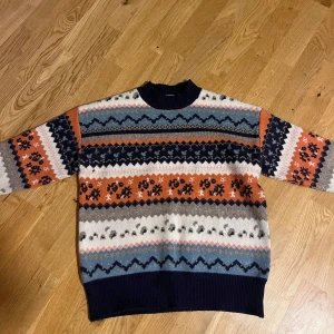 Mönstrad stickad tröja i retrostil - Fet stickad tröja med retrovibbar och coolt mönster i blått, orange, vitt, grått och marinblått. Tröjan har rund hals, långa ärmar och breda muddar. Perfekt för dig som gillar färg nu i höst och vinter! Passar xs och s. Nytt skick!