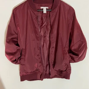 Vinröd tunn bomberjacka från Nelly  - Snygg vinröd bomberjacka från Nelly med klassisk ribbad krage, muddar och dragkedja framtill. Jackan är tillverkad i glansig polyester och har två fickor på sidorna. Perfekt för dig som vill ha en stilren och enkel look. 