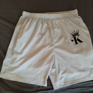 Vita shorts med K-logga och krona - Snygga vita shorts med elastisk midja och svart K-logga med krona på vänster ben. Perfekta för sommaren och chill dagar. Lätta och bekväma med sportig vibe, tillverkade i mjukt syntetmaterial som andas.