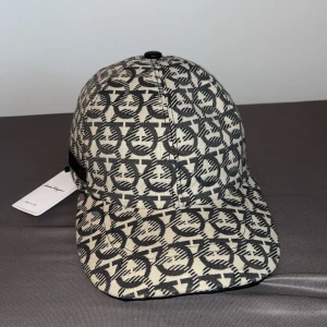Salvatore Ferragamo mönstrad keps - Snygg keps från Salvatore Ferragamo med svart och beige grafiskt mönster över hela kepsen. Justerbar rem baktill med metallspänne. Tillverkad i bomull och har en klassisk böjd skärm. Perfekt för dig som vill sticka ut med designerstil. Ferrargamo keps ny använd 3 gånger max inga skador eller hål eller något sånt tar byten och pris kan diskuteras vid nabb affär