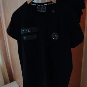 Svart Philipp Plein t-shirt med logga - Svart t-shirt från Philipp Plein med coola detaljer som två svarta tygpatches med silvrig text och en sexkantig logga på bröstet. Klassisk rund hals och korta ärmar. Snygg och stilren design som sticker ut. (Passar till M mer än L)