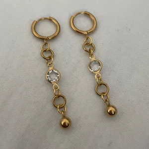 Guldiga hängande örhängen med kristall - Snygga guldiga örhängen med hängande kedjor, runda länkar och en klar kristall i mitten. Längst ner sitter en liten guldfärgad kula som ger extra bling. Perfekt för dig som gillar statement-smycken och vill sticka ut. Rostfritt stål. 