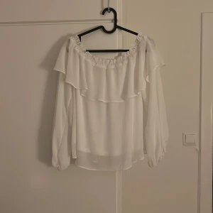 Vit offshoulder blus med volang - Superfin vit blus med offshoulder-modell och bred volang runt hela överdelen. Långa, luftiga ärmar med resår vid handlederna ger en romantisk vibe. Perfekt för dig som gillar en luftig och flowy stil.