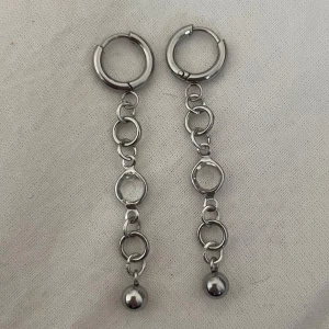 Silvriga kedjeörhängen med kula - Säljer ett par coola örhängen i silverfärgad metall med hängande kedjor och en liten rund kula längst ner. Designen är modern med flera ringar som ger en edgy vibe. Perfekt för dig som gillar statement-accessoarer och vill sticka ut. Rostfritt stål. 