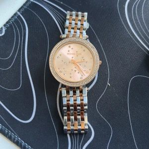 Glittrig klocka från Michael Kors - Snygg klocka från Michael Kors med glittrande stenar och stjärnor på urtavlan. Armbandet är i silver och roséguld med en klassisk länkdesign. Runt urtavlan sitter en rad med små glittrande stenar som ger extra bling. Perfekt accessoar för att lyfta din stil.