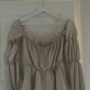 Beige puffärm topp & volang - Supersöt beige topp med puffiga långa ärmar och volang vid halsringningen. Klänningen har offshoulder-look och är gjord i ett mjukt, glansigt material💕står ingen storlek men gissar på S!