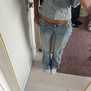 Ljusblå bootcut jeans med fransar - Säljer dessa eftertraktade ljusblåa jeanen med bootcut passform och fransade detaljer längs sidorna. Byxorna har låg midja och klassisk femficksdesign. Perfekta för dig som gillar en chill och trendig stil med lite edge. Materialet är jeans och färgen är ljusblå.