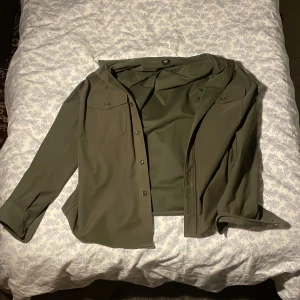 Olivgrön skjorta från Uniqlo, M - Säljer en stilren olivgrön skjorta från Uniqlo i storlek M. Skjortan har klassisk krage, knappar framtill och två bröstfickor med lock. Perfekt för dig som gillar minimalistisk och clean stil.