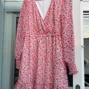 Blommig kortklänning från Amisu - Superfin kortklänning från Amisu med rosa och röda blommor på vit botten. Klänningen har omlott-v-ringad front, långärmade ärmar med smock vid mudden och volangdetalj längst ner. Perfekt för sommarens alla tillfällen!