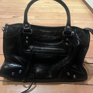 Svart handväska - Snygg svart handväska med silverfärgade detaljer och dragkedja framtill. Väskan har dubbla handtag, innerfack med dragkedja och flera småfack för smart förvaring. Perfekt storlek för att få plats med allt du behöver.