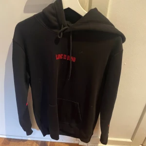 Svart hoodie med broderade rosor - Svart hoodie med röd text 'LOVE IS BLIND' på bröstet och broderade röda rosor längs båda ärmarna. Tröjan har huva med snörning och en stor magficka framtill. Perfekt för dig som gillar unika detaljer och streetstyle.