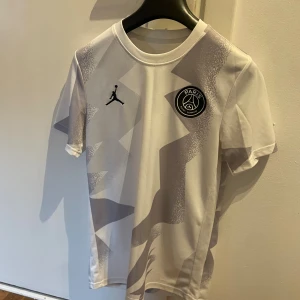 PSG x Jordan vit fotbollströja - Snygg vit PSG fotbollströja i samarbete med Jordan. Tröjan har korta ärmar, coolt geometriskt mönster i grått och PSG-logga på bröstet. Tillverkad i lätt och ventilerande material som passar perfekt till träning eller match.