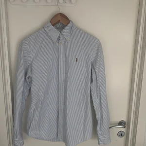 Randig skjorta från Ralph Lauren - Klassisk skjorta från Ralph Lauren i ljusblått och vitt med smala ränder. Skjortan har button-down krage, broderad logga på bröstet och långa ärmar med manschettknappar. Perfekt för en clean och stilren look. Hör av er vid funderingar 