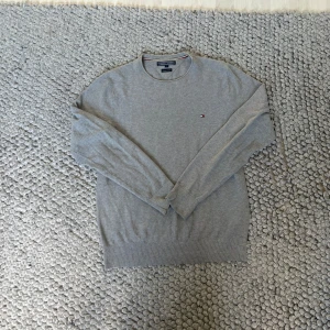 Grå Tommy Hilfiger tröja - Säljer en stilren grå tröja från Tommy Hilfiger i premium bomull. Klassisk rund halsringning, diskret logga broderad på bröstet och ribbade muddar. Perfekt för dig som gillar enkel och clean stil. 67,5cm från axel och 48cm bred