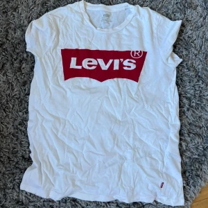 Vit Levi's t-shirt med rött tryck - Vit t-shirt från Levi's med det klassiska röda logotrycket framtill. Modellen är kortärmad och har rund hals. Materialet är mjuk bomull som känns skönt mot huden. Perfekt för dig som gillar ikoniska streetwear-plagg och enkel stil.