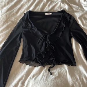 Svart omlottopp med volang från Nelly - Jättesnygg topp från Nelly med knytning i fram. Säljer pågrund av att den inte kommer till användning. 