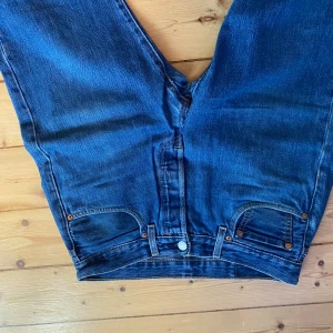 Levi's 501 blå jeans straight fit - Klassiska Levi's 501 jeans i blå denim med raka ben och normal passform. Byxorna har fem fickor, knappgylf och tydliga kontrastsömmar. Ikonisk läderpatch bak i midjan och röd Levi's-tagg på bakfickan. Perfekta för en avslappnad och tidlös stil.