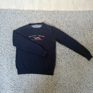 Mörkblå stickad tröja från Gant - Snygg mörkblå stickad tröja från Gant med broderad logga och amerikansk flagga på bröstet. Klassisk rund halsringning och långa ärmar. Perfekt till jeans för en clean och sportig look. 67cm från axel och 49cm bred 