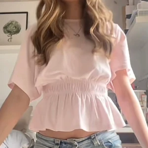 Ljusrosa topp från lager 157 - Supersöt ljusrosa topp från lager 157 med smockad midja och volang nedtill. Toppen har korta, vida ärmar och är croppad för en trendig look. Perfekt att matcha med jeans eller kjol för en chill och söt stil. Endast använd 1 gång!🩷🩷